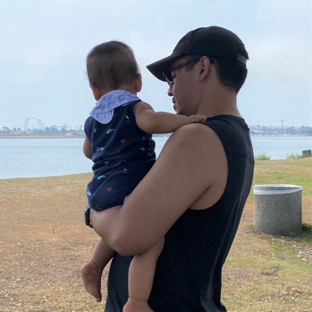 Me standing at the bay holding my son in my left arm. My left shoulder is facing the camera and so is his back. He's wearing a blue tank top and a white bib. I'm wearing a black hat and a black sleeveless shirt.
